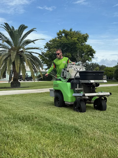 Glade Palm technician applying a lawn treatment with a ride-on spreader beside a palm tree